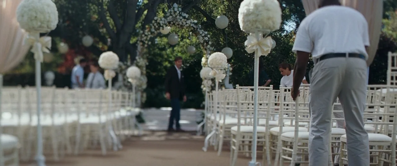 The Hangover (2009) movie still: medium — An outdoor wedding venue features rows of white chairs leading toward a floral arch. A man in a white polo…