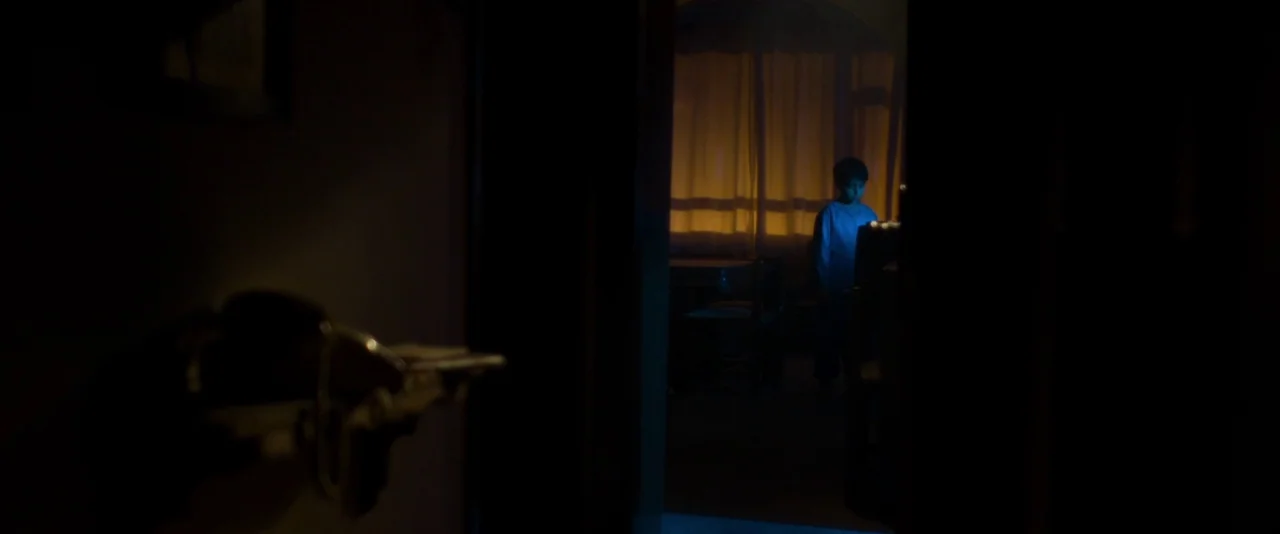 Baskin (2015) movie still: wide — A dark, cluttered room is viewed through a partially opened doorway. A child stands illuminated by blue light in…