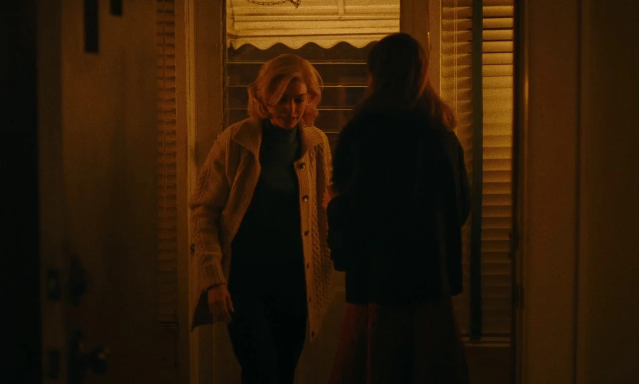 Eileen (2023) movie still: medium — A dimly lit residential threshold is framed by a doorway with venetian blinds. A blonde woman enters from…