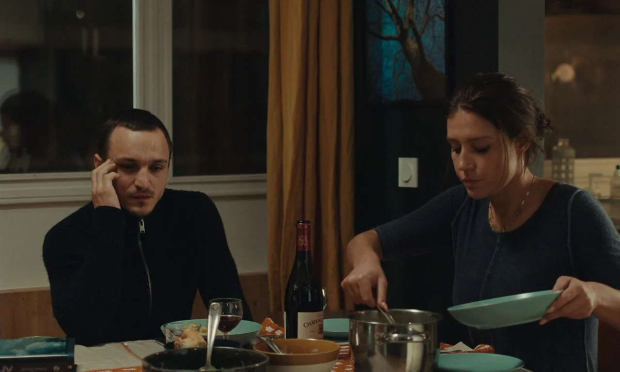 Passages (2023) movie still: medium — A modern dining room features a table set with wine, bowls, and a metal pot. A man sits quietly watching while…