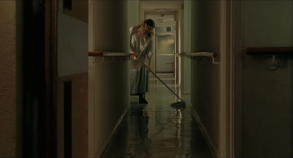 All or Nothing (2002) movie still: wide — A long, dimly lit institutional corridor has pale green walls and a wet tiled floor. A woman in a white…