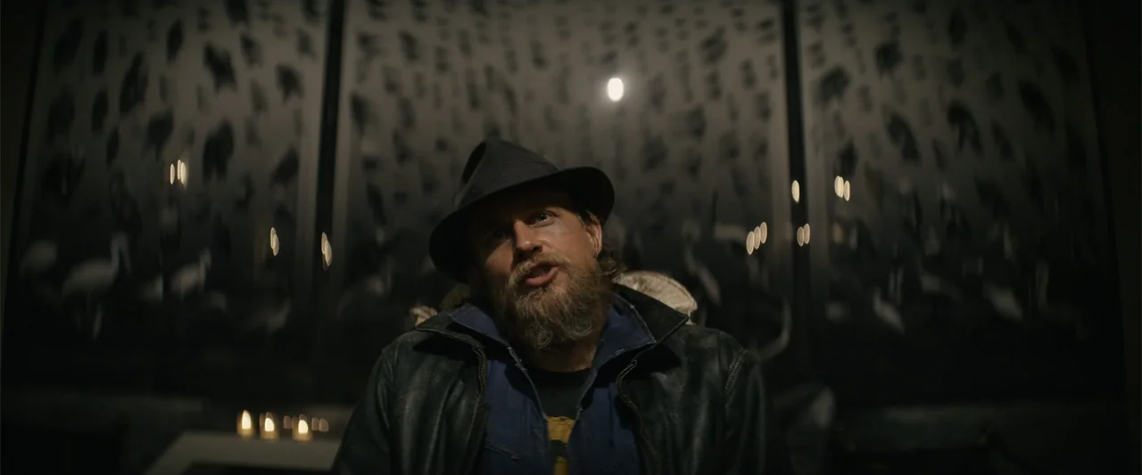 Last Looks (2022) movie still: medium — A dark lounge features wall-mounted abstract art panels illuminated by low flickering light. A man in a…