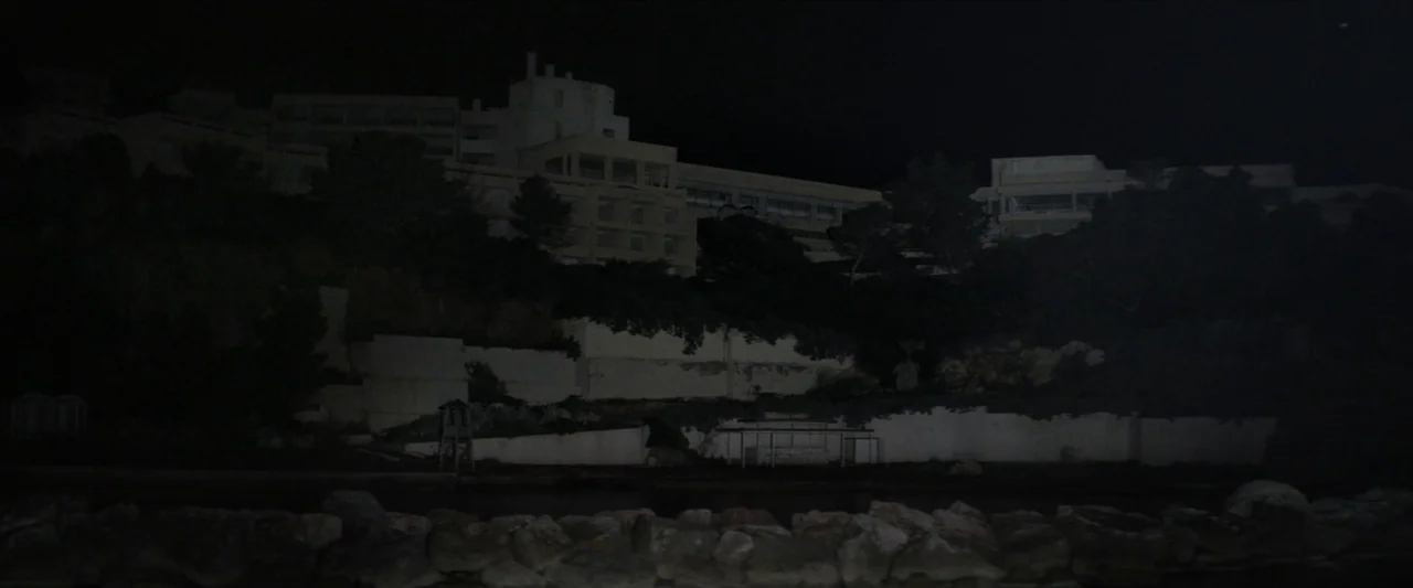 Chevalier (2015) movie still: establishing — A dark, multi-story hotel complex sits atop a rocky, overgrown cliffside at night. The structure is…