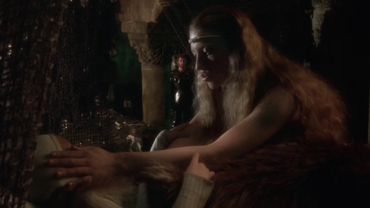 Excalibur (1981) movie still: medium — A stone chamber with arched architecture is draped with dark fishing nets and fur blankets. A woman with long…