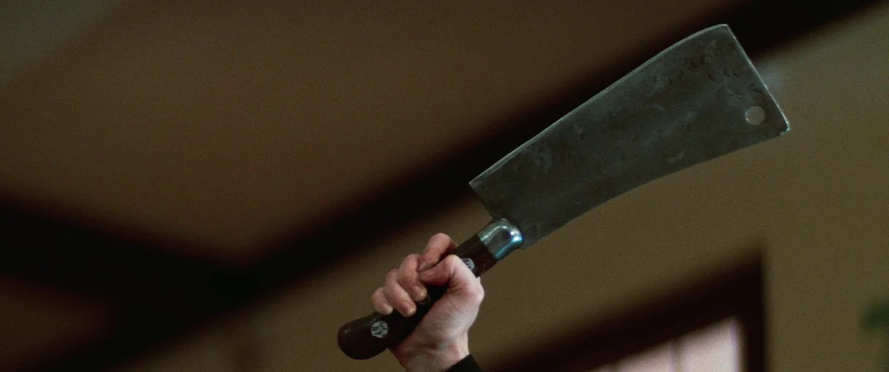 Death Game (1977) movie still: closeup — A dark indoor space with blurred background walls and a window frame. A person's hand grips a large, worn…