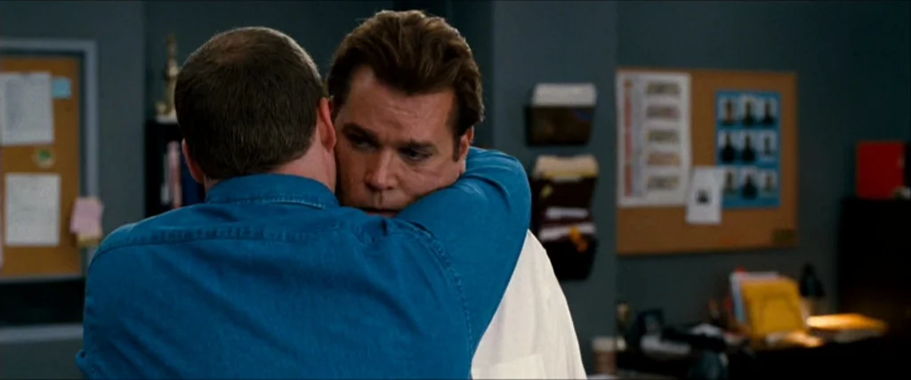Observe And Report (2009) movie still: medium — An office area with grey walls features a cork board with pinned papers. A man in a blue shirt hugs…