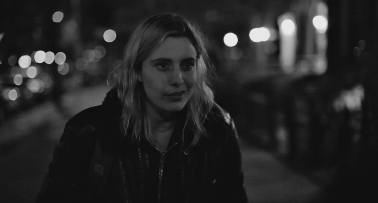Frances Ha (2012) movie still: medium — A nighttime city street serves as the setting with bokeh lights in the background. A woman in a leather…