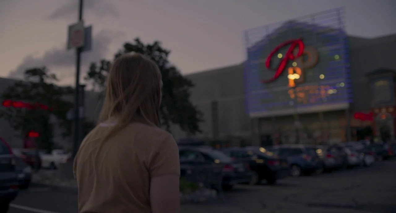 Eighth Grade (2018) movie still: medium — A view across a populated parking lot toward a large commercial structure at dusk. A young woman walks…