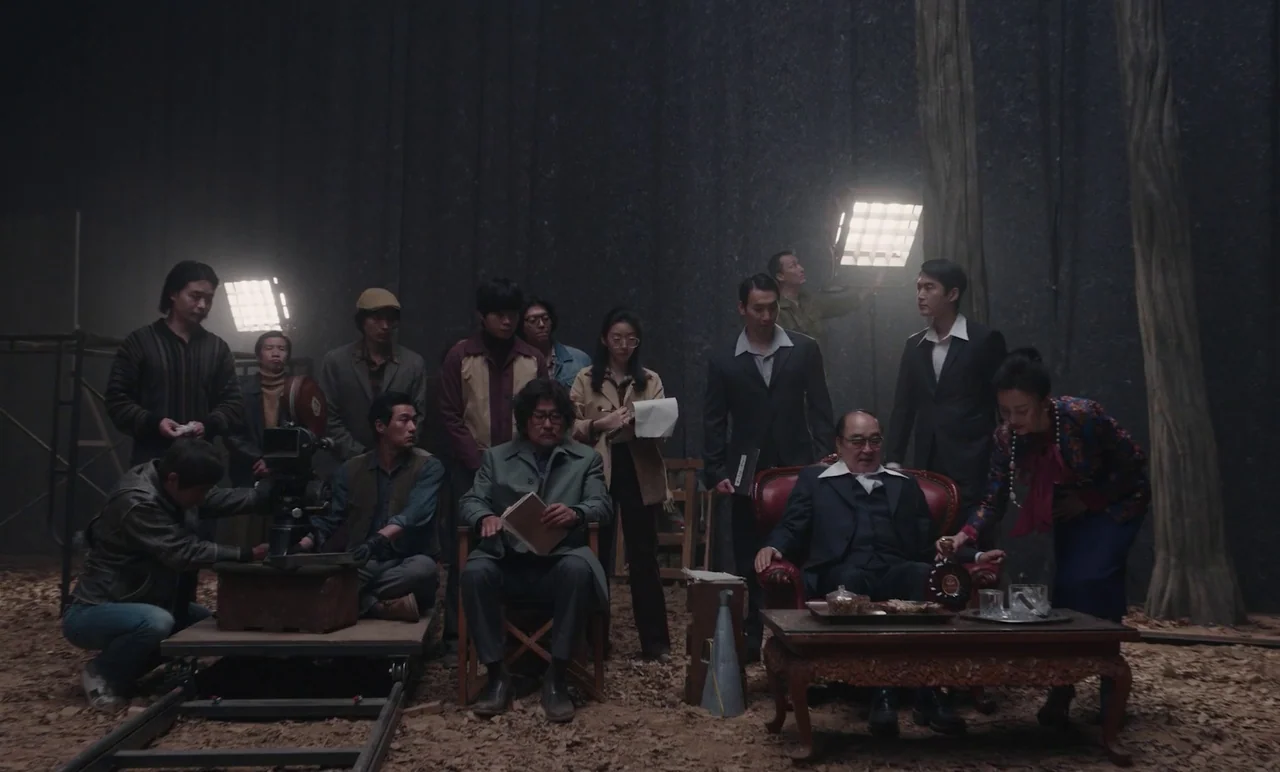 Cobweb (거미집) (2023) movie still: wide — A dark film set interior with scattered wood shavings on the floor surface. A large production crew observes…