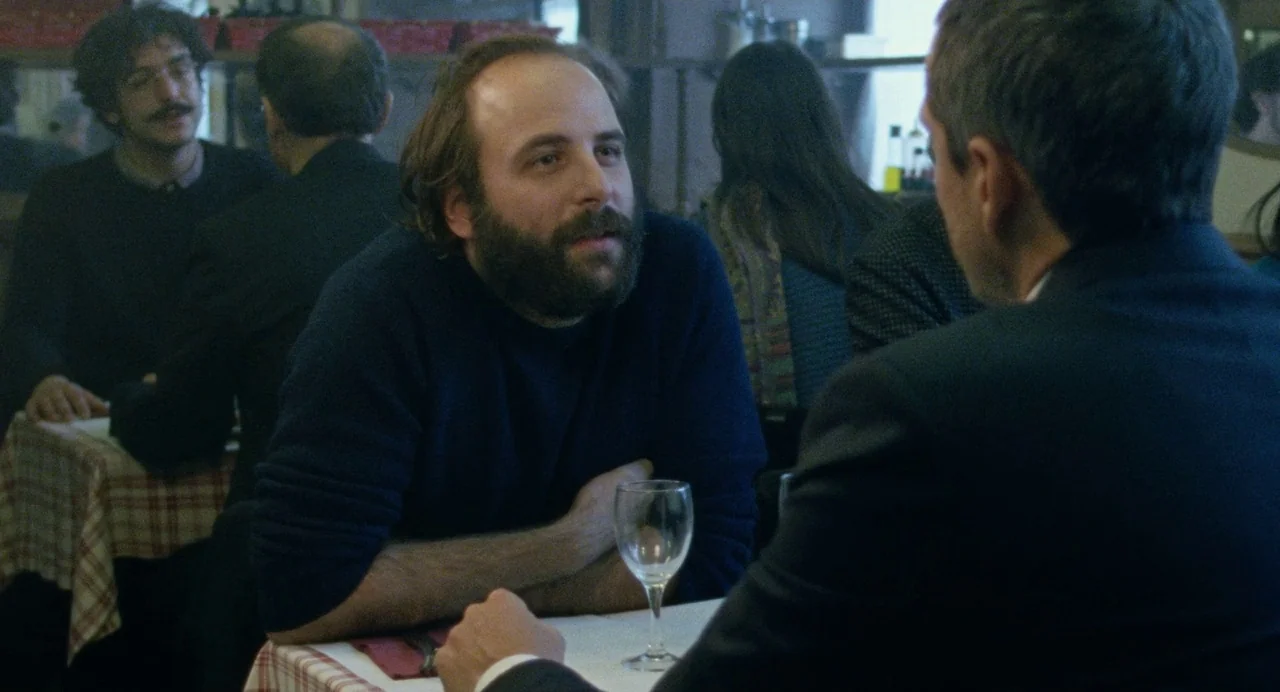Non-Fiction (2018) movie still: medium — A quiet bistro interior features tables with checkered tablecloths and multiple patrons seated. A bearded…