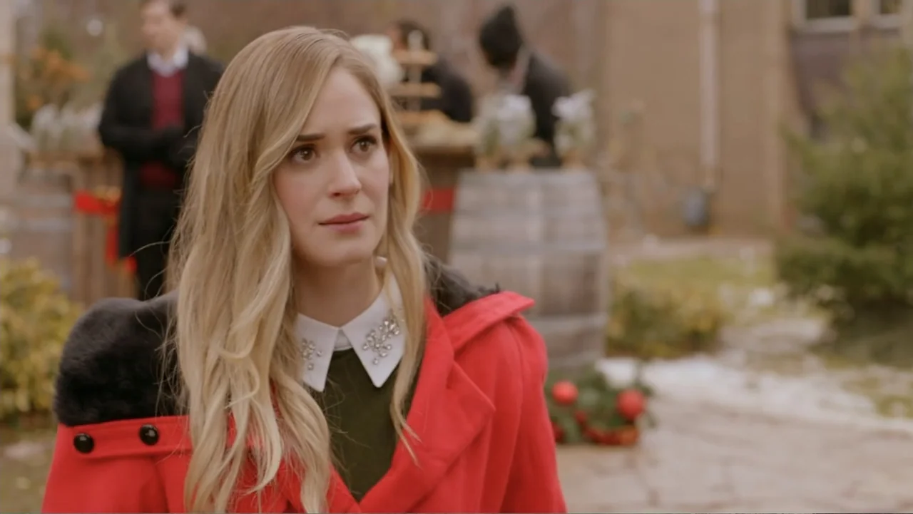 Loving Christmas (2022) movie still: medium — An outdoor courtyard area features a wooden barrel and blurred figures in the background. A woman in a…