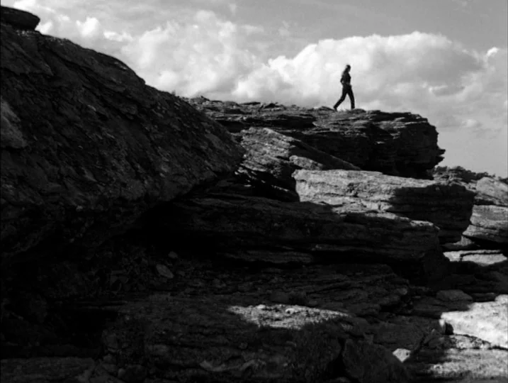 Persona (1966) movie still: wide — A rugged terrain features expansive layered rock formations under a cloudy sky. A solitary person walks along the…