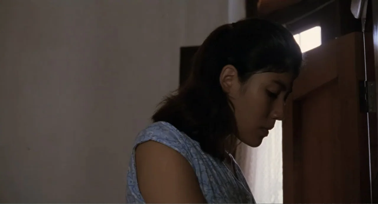 La Ciénaga (2001) movie still: medium — A dimly lit room interior features a plain wall and an open doorway. A young woman with dark hair in a…