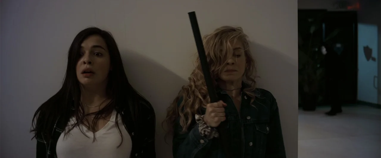 Init!ation (2021) movie still: medium — A stark white interior wall serves as the backdrop for two women in a hallway. The women stand…