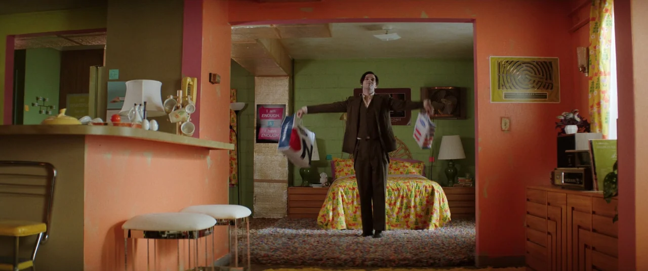 Renfield (2023) movie still: fullbody — An apartment interior features vibrant orange walls and a green-walled sleeping area beyond. A man in a…