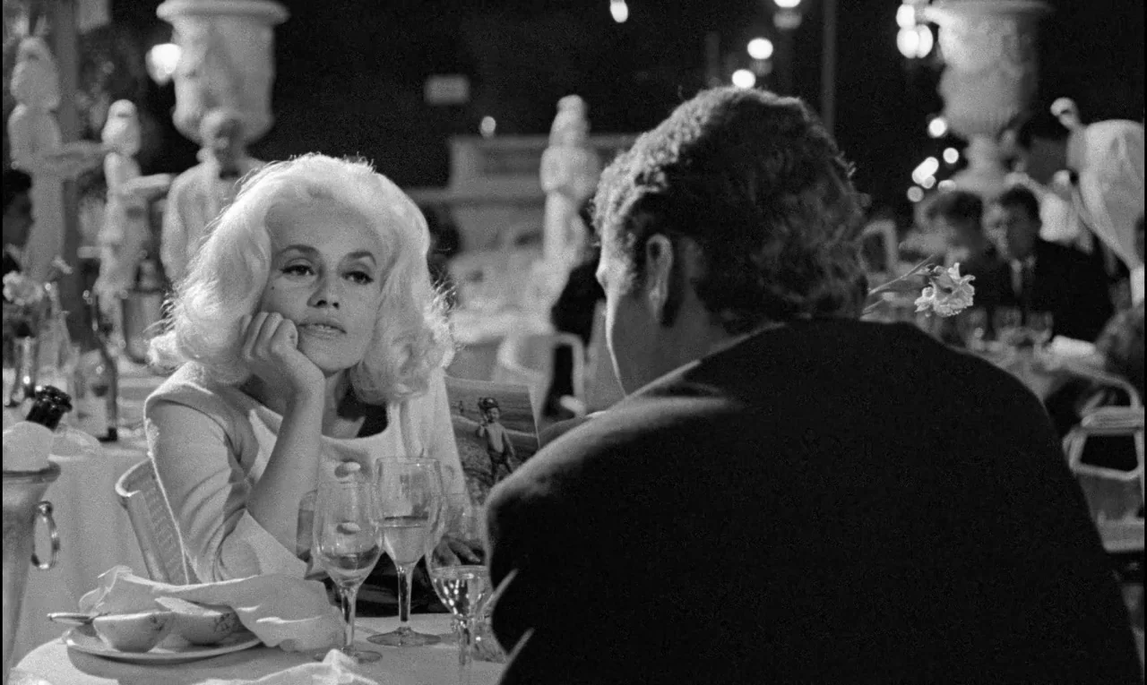 Bay of Angels (1963) movie still: medium — An outdoor dining area at night features white-clothed tables and ornate stone urns. A blonde woman sits…