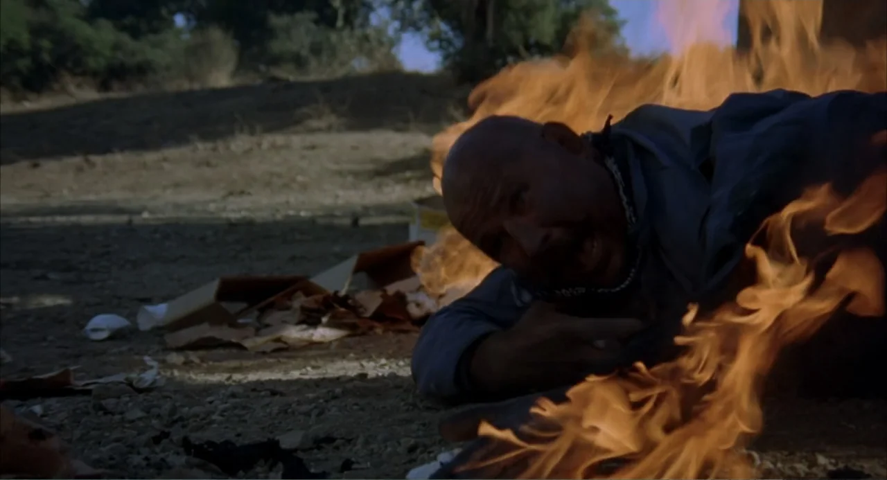 Foxy Brown (1974) movie still: medium — A dry, sunlit desert clearing features scattered debris on the parched earth floor. A bald man lies face…
