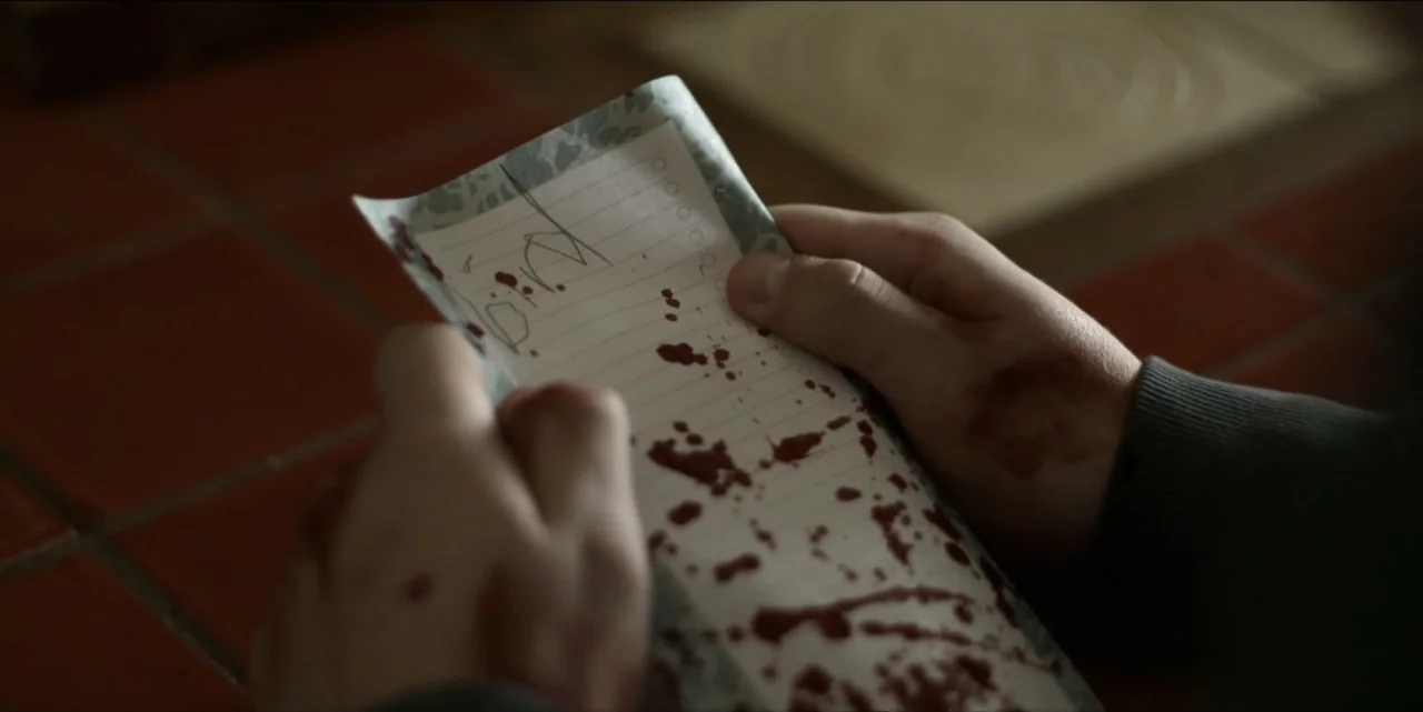 Bring Her Back (2025) movie still: closeup — A close view shows two hands holding a small piece of lined notebook paper. The paper is covered in red…
