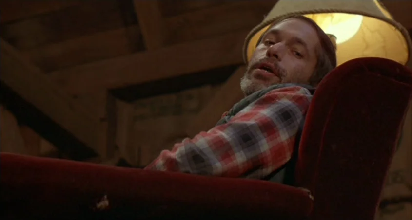 Scanners (1981) movie still: medium — A dimly lit interior space with wooden ceiling beams and a large armchair. A man with a beard sits in the…