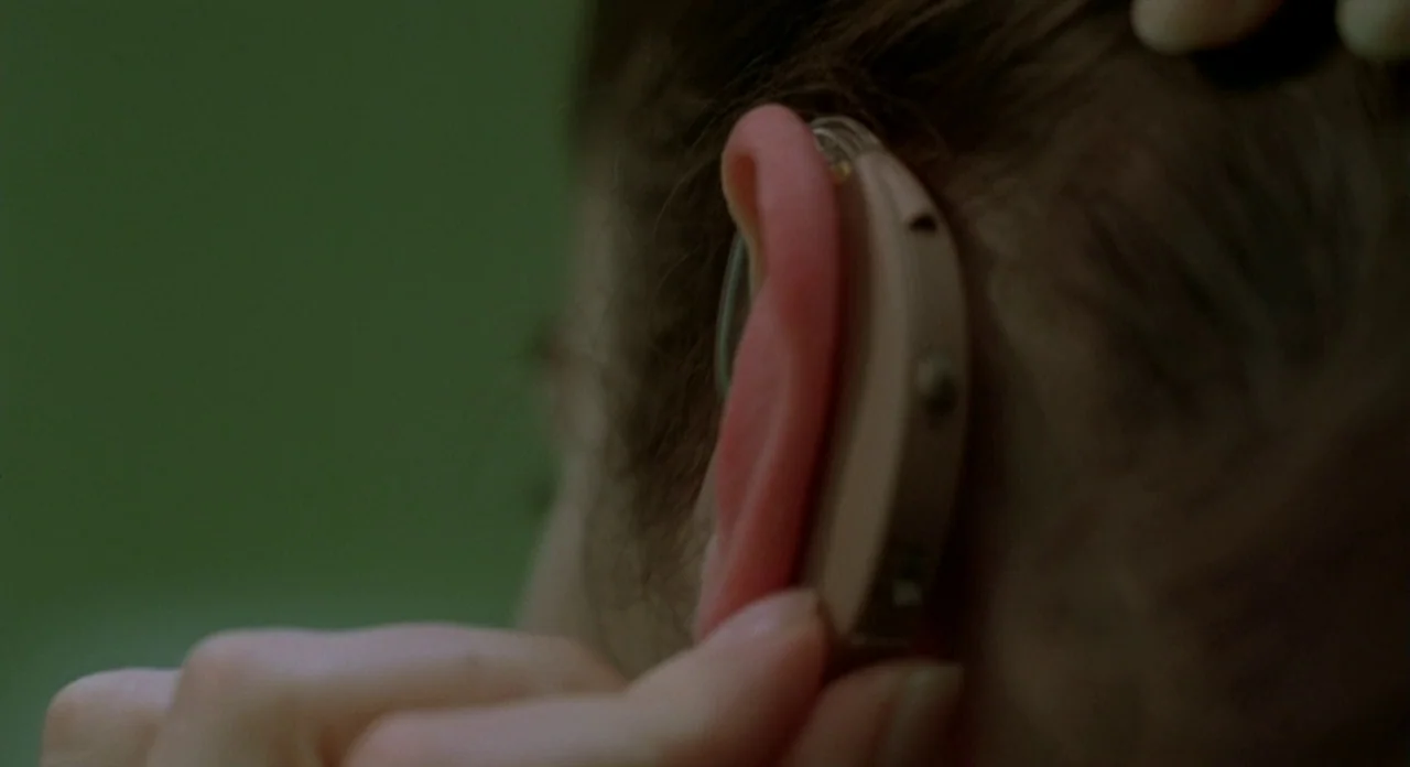 Read My Lips (2001) movie still: closeup — A close-up view focuses on a human ear against an indistinct olive green backdrop. A hand gently touches…
