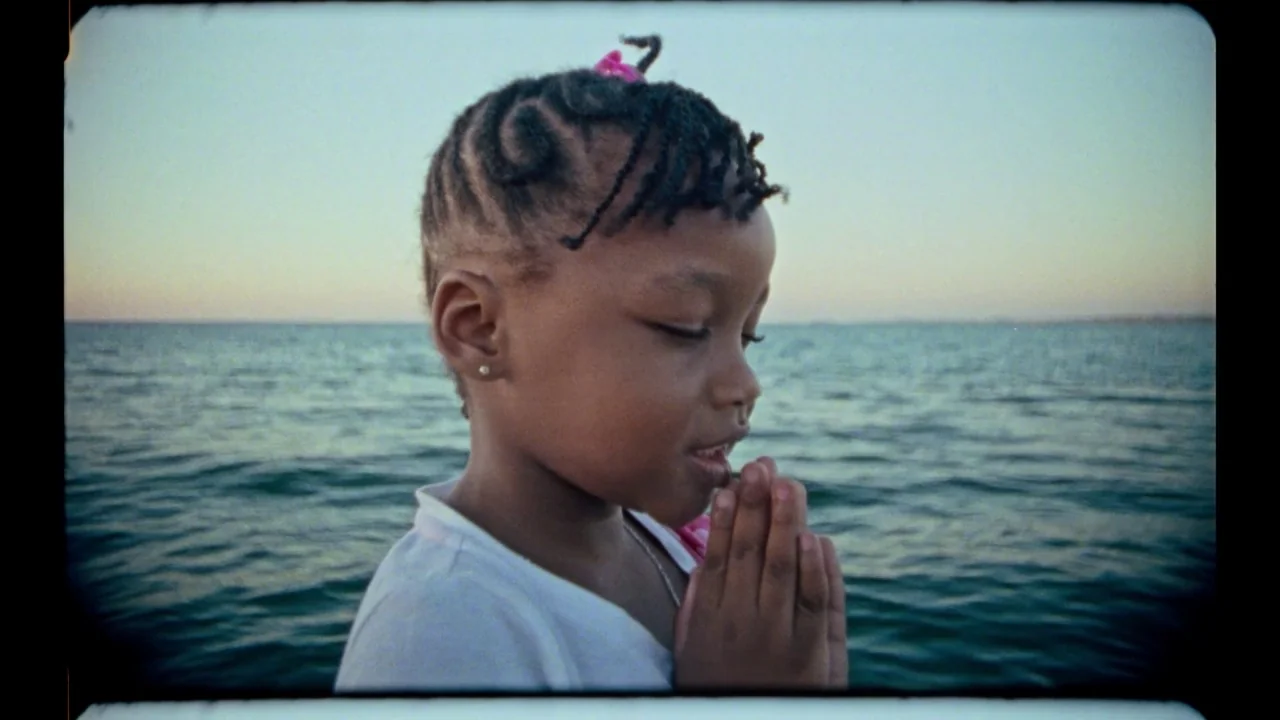 Black Mother (2018) movie still: closeup — A young child stands in front of the open sea during golden hour. She holds her hands together in a…