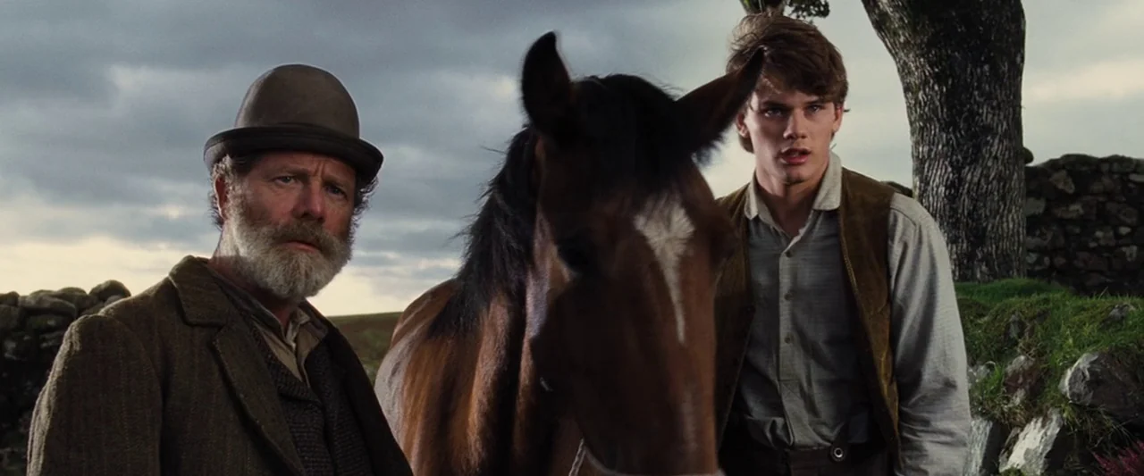 War Horse (2011) movie still: medium — A man and a youth stand with a horse in a rural open landscape. They are framed against a cloudy sky with dry…