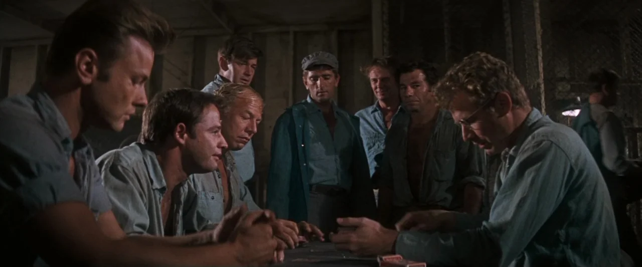 Cool Hand Luke (1967) movie still: medium — A dimly lit room with wooden walls and wire mesh partitions houses several men. The group sits around a…