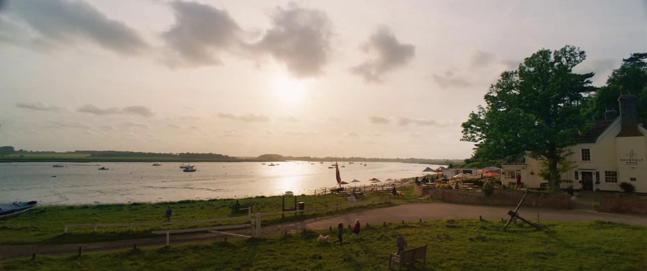 Yesterday (2019) movie still: wide — A riverside pub stands next to a wide tidal waterway at golden hour. Several small children run and walk across…