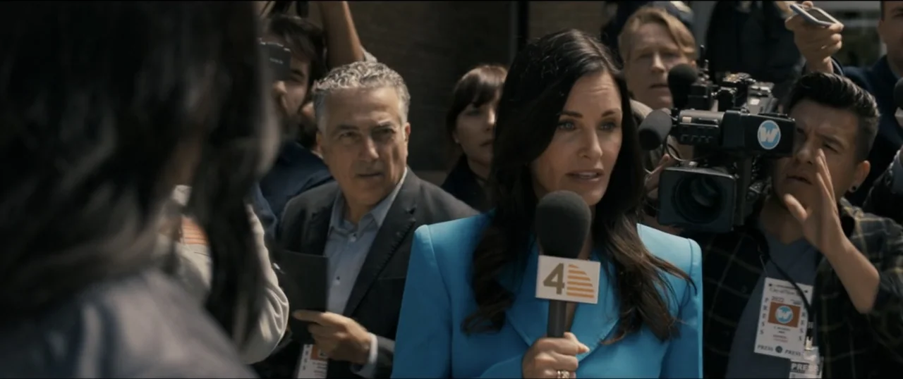 Scream VI (2023) movie still: medium — A group of reporters and cameras surrounds a news anchor outdoors. The woman in the foreground speaks into a…