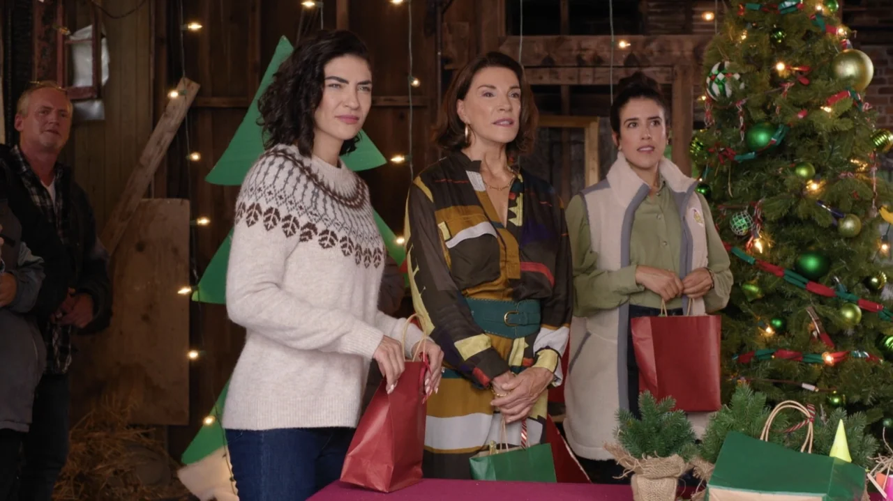 Designing Christmas (2022) movie still: medium — A rustic barn interior decorated for Christmas features a lit tree and string lights. Three women…
