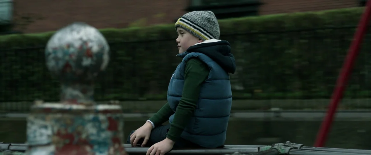 The Cured (2017) movie still: medium — A child sits on a dark metal railing along a canal. A mossy brick wall runs horizontally behind him under an…