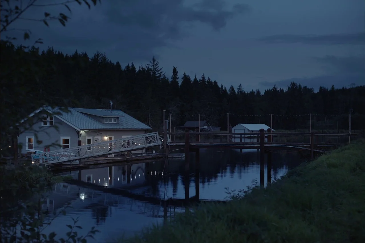 Sometimes I Think About Dying (2023) movie still: establishing — A wooden dock extends from a grassy bank toward a floating structure at dusk. Dark…