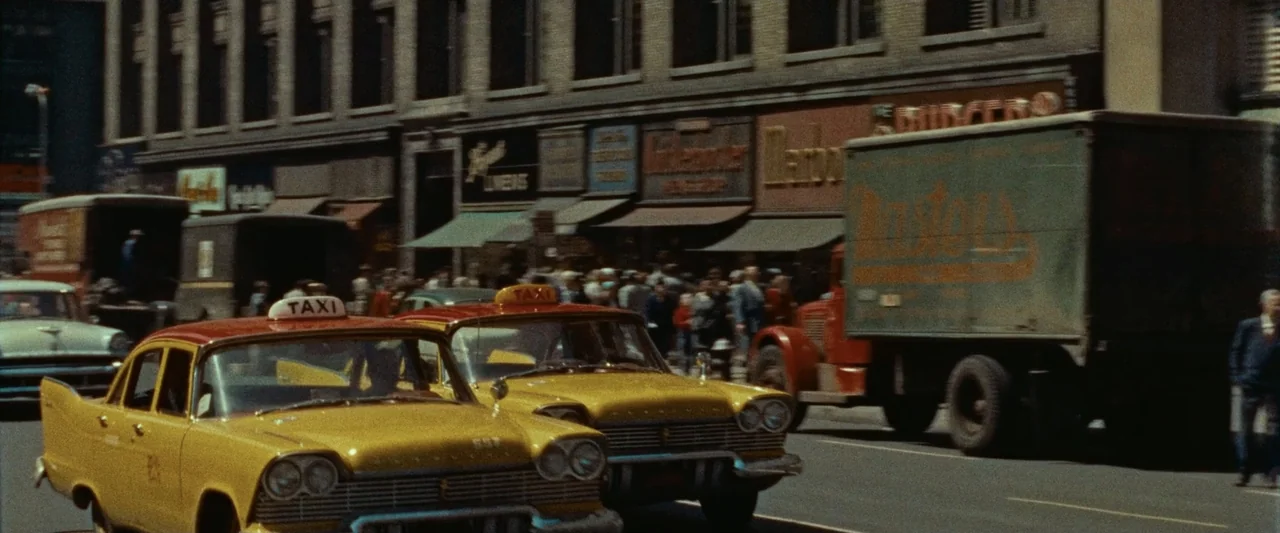The Alto Knights (2025) movie still: wide — A bustling 1950s city street with multiple storefronts and vintage signage in the background. Two yellow…
