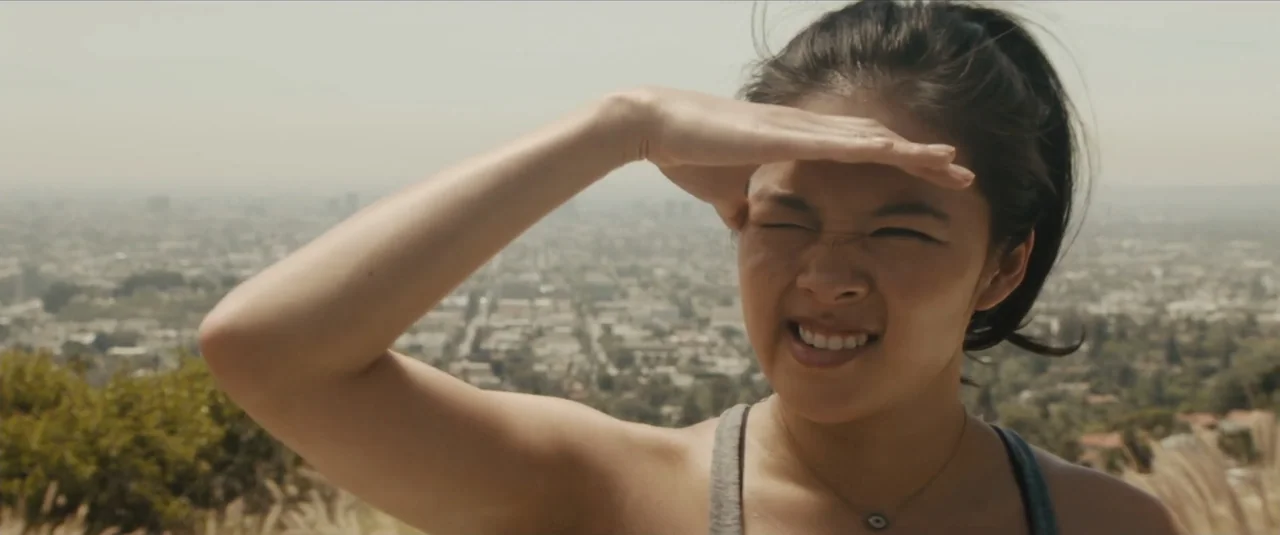 Stalker (2020) movie still: closeup — A hillside overlook provides a panoramic view of a distant, hazy urban sprawl. A young woman in a grey tank…