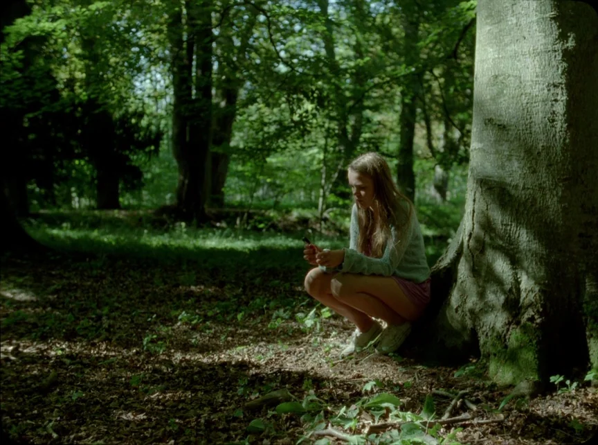 Jauja (2014) movie still: fullbody — A teenage girl crouches at the base of a large, moss-covered tree in a forest. She holds a small dark object in…