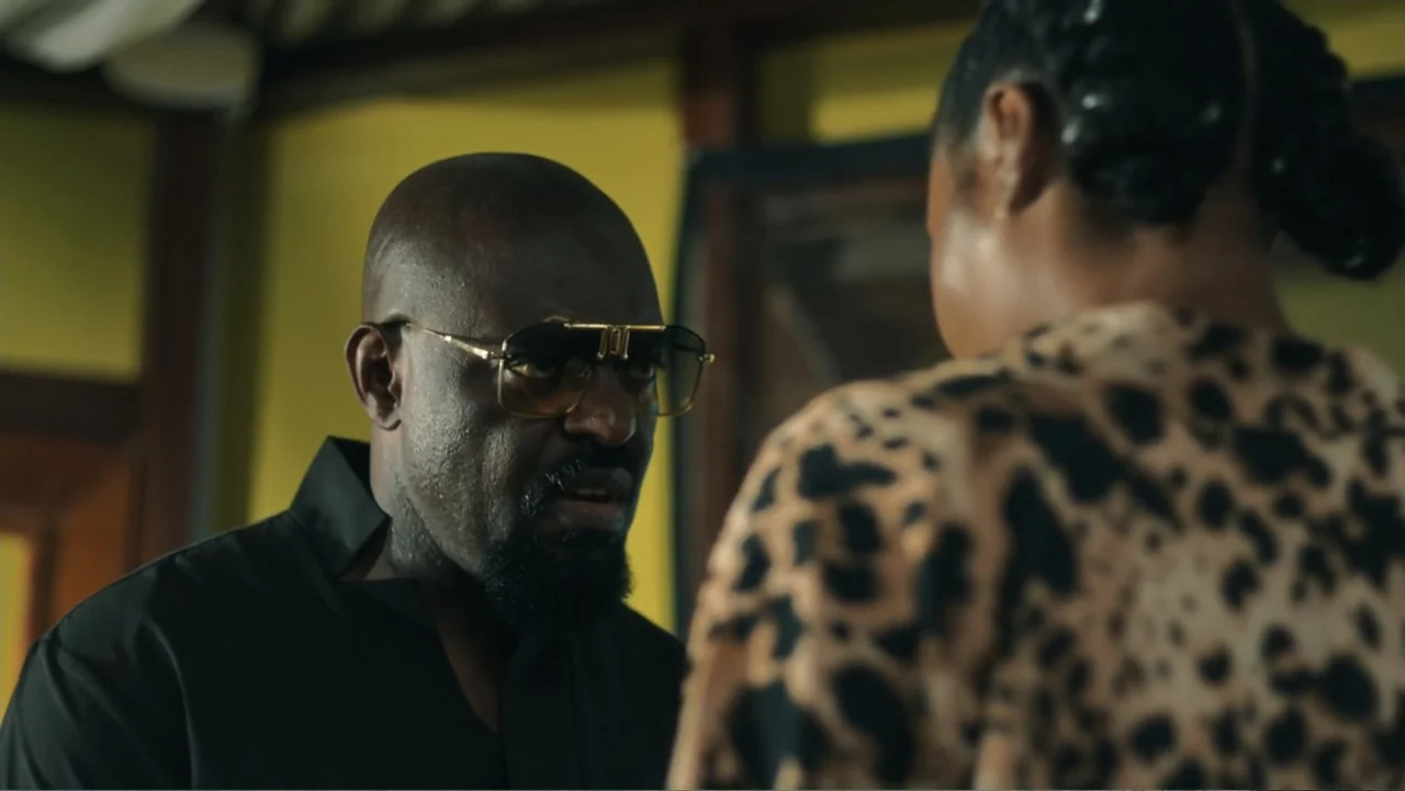 SIN: Blackout (2025) movie still: medium — An interior office space features a yellow wall and dark wooden furniture elements. A bald man wearing…