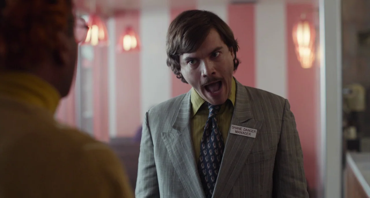 An Evening With Beverly Luff Linn (2018) movie still: medium — A man with a name tag stands in a diner with striped wallpaper. He is yelling at…