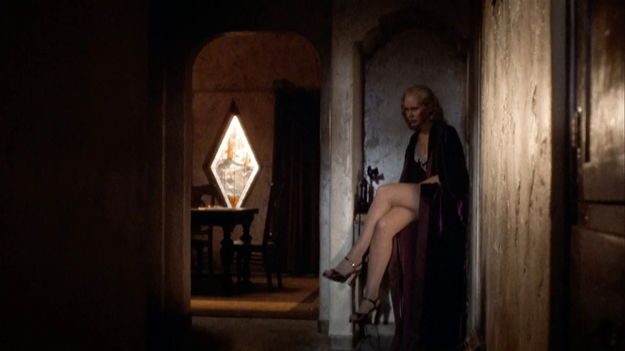 The Day of the Locust (1975) movie still: medium — A dimly lit narrow hallway with textured walls and an arched doorway. A woman sits on a small…