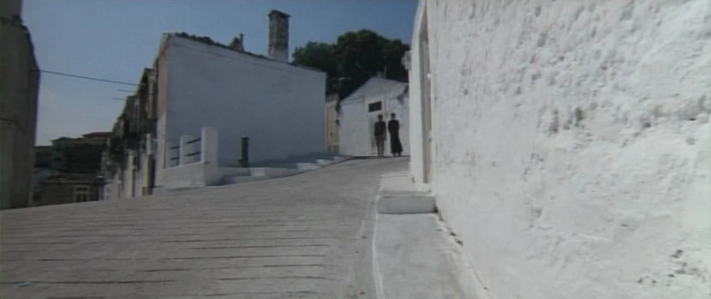 Don’t Torture A Duckling (1972) movie still: wide — A narrow, stone-paved path bordered by stark white, lime-washed Mediterranean buildings rises…