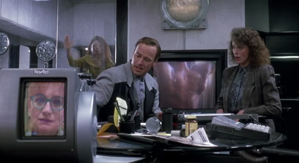 Total Recall (1990) movie still: medium — A high-tech office desk is crowded with computers and assorted office supplies. A man and a woman analyze…