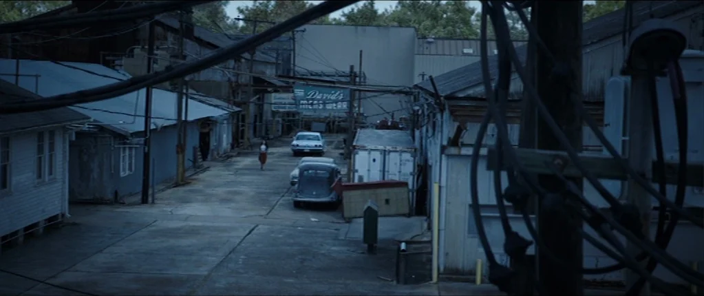 Abattoir (2016) movie still: wide — A narrow service alley between industrial buildings features overhead power lines and parked vintage cars. A…