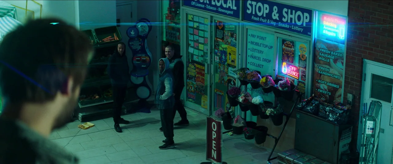 Unwelcome (2022) movie still: wide — The exterior of a small city shop with glass windows and a neon sign. Three young men stand outside the…