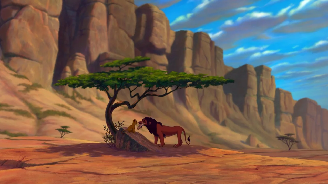 The Lion King (1994) movie still: wide — A wide desert savanna features steep rocky cliffs under a bright blue sky. An adult lion and a cub interact…