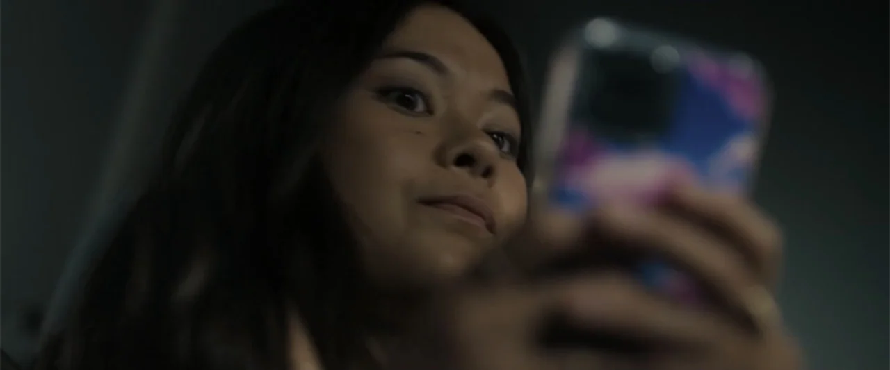 Afraid (2024) movie still: closeup — A dark indoor space with minimal ambient light cast by a handheld screen. A young woman with long dark hair…