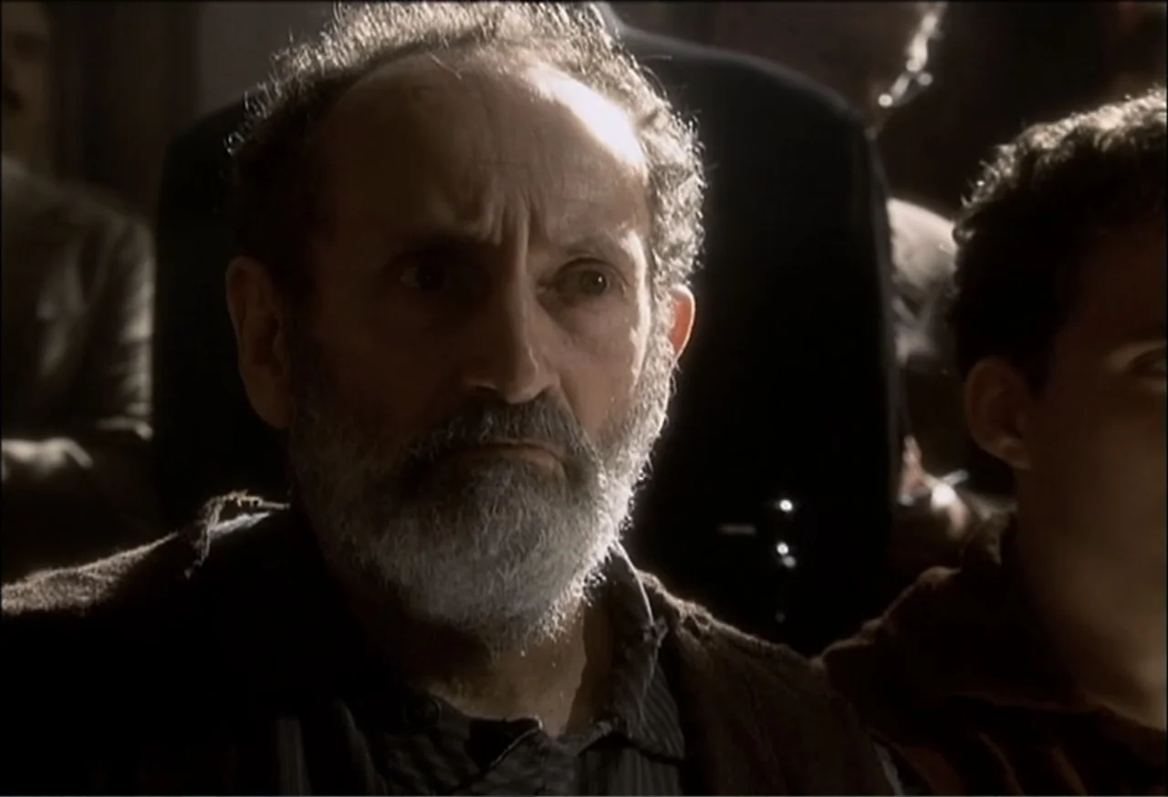 Vendetta (2022) movie still: closeup — A dark, crowded interior space with figures visible in the dimly lit background. An elderly man with a grey…