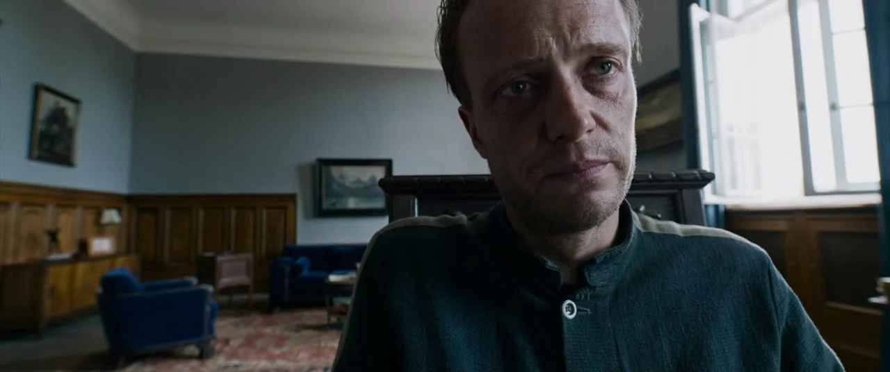 A Hidden Life (2019) movie still: closeup — A study with blue walls, wood paneling, and period furniture including chairs and paintings. A man sits…