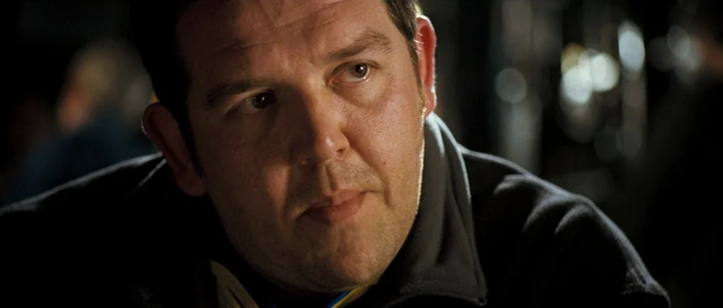 Hot Fuzz (2007) movie still: closeup — A dimly lit interior space with an indistinct, dark background. A man sits, facing slightly away from the…