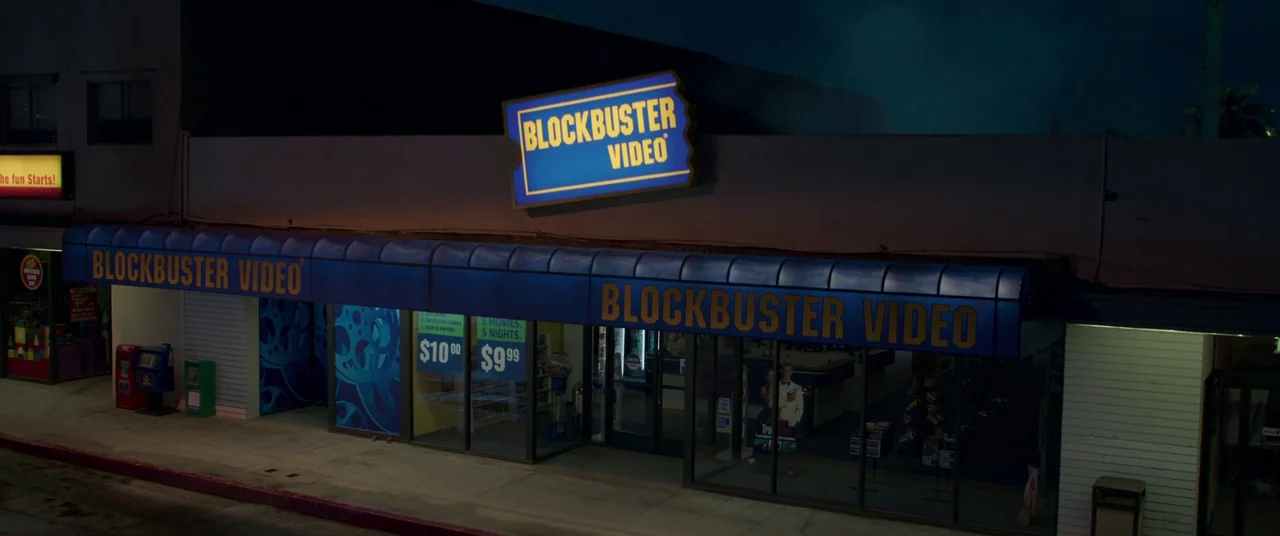 Captain Marvel (2019) movie still: wide — A night-time view of a Blockbuster Video retail storefront with illuminated blue signage above. The glass…