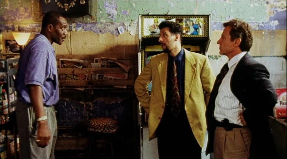 Clockers (1995) movie still: medium — A convenience store interior features peeling wallpaper and an arcade cabinet against the wall. Three men…