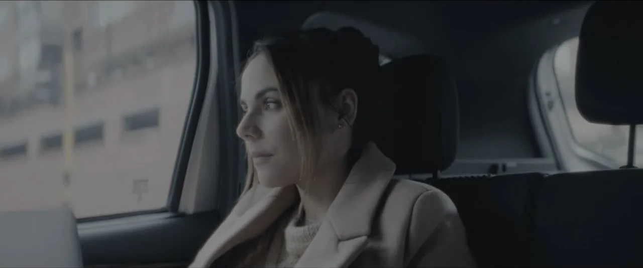 Quicksand (2023) movie still: medium — The interior of a car backseat shows a woman looking toward a window. Outside the window, a blurred urban…